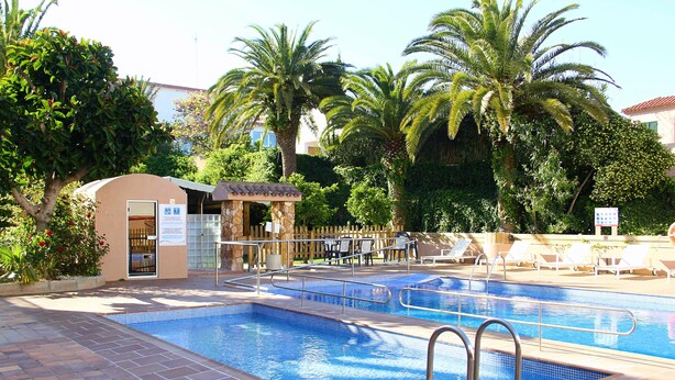 A swimming pool with a tiled deck, lounge chairs, and palm trees. - Apartamentos Royal Life (Mahón)