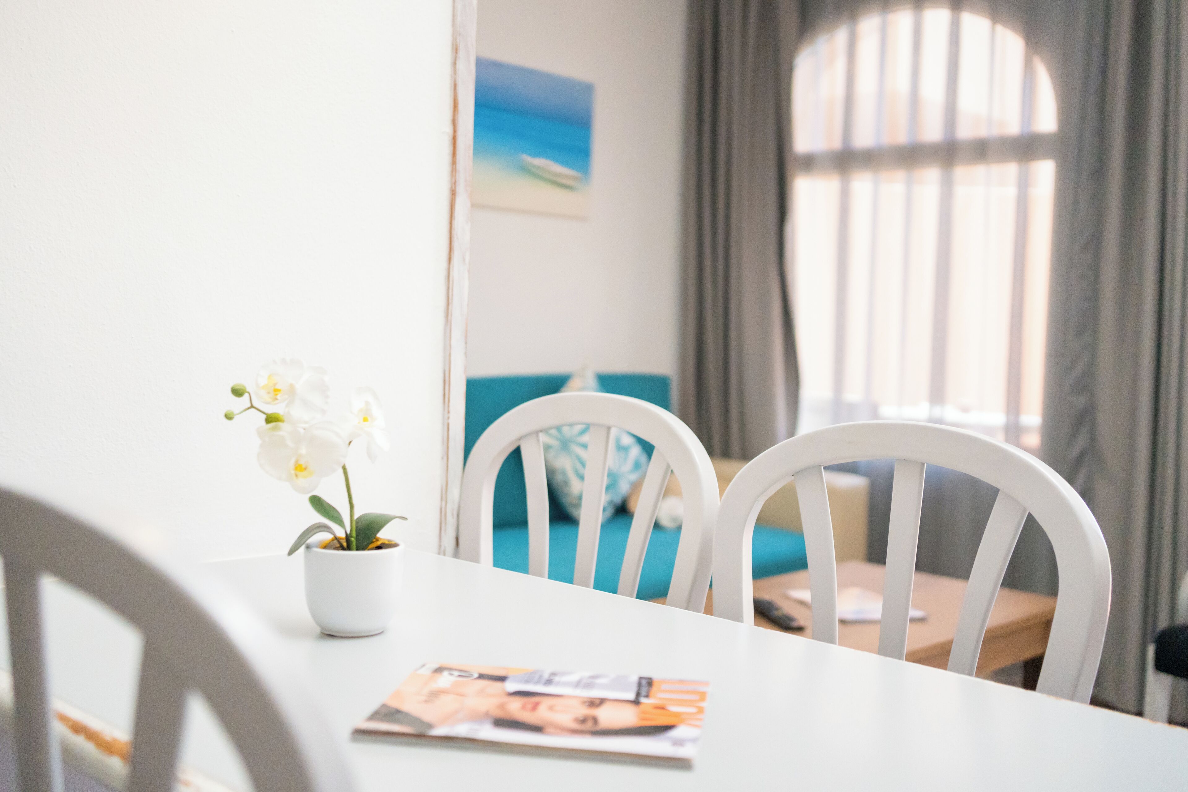 A dining table with a magazine, a potted orchid, and white chairs.