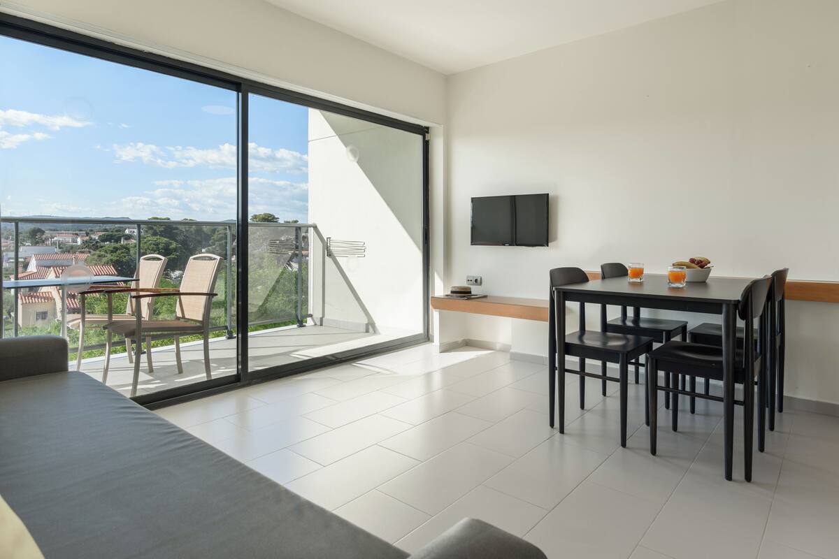 A modern living room with a sofa, a dining table with chairs, a TV mounted on the wall, and a balcony with a view of the city.