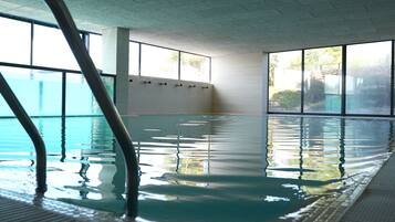 An indoor swimming pool with clear water, large windows, and a tiled wall.