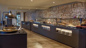 A buffet area with a stone wall, multiple food stations, and a coffee machine. - FERGUS Cap Roig (L'Ampolla)