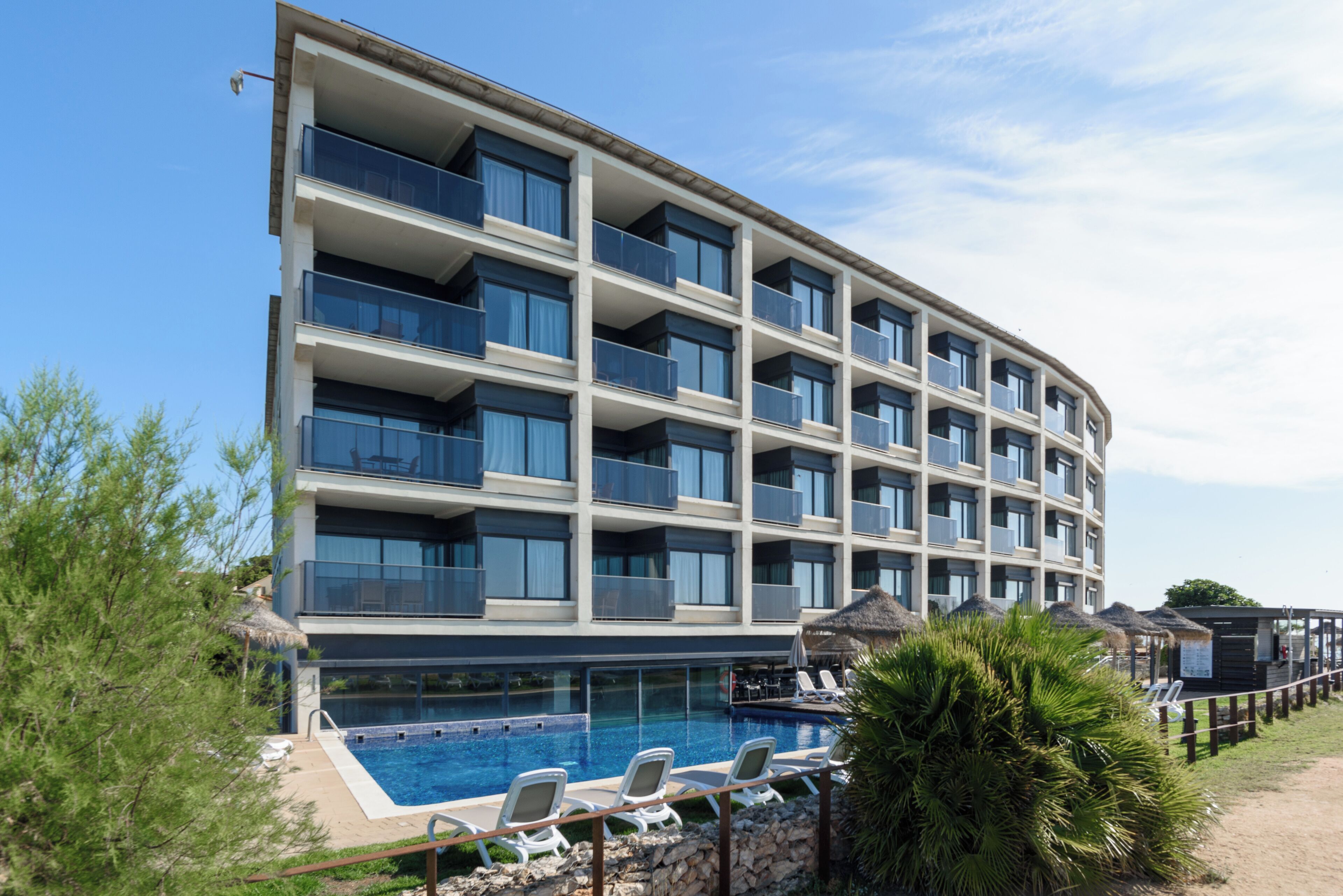 Un hotel de varios pisos con piscina, mobiliario exterior y una estructura de techo de paja.