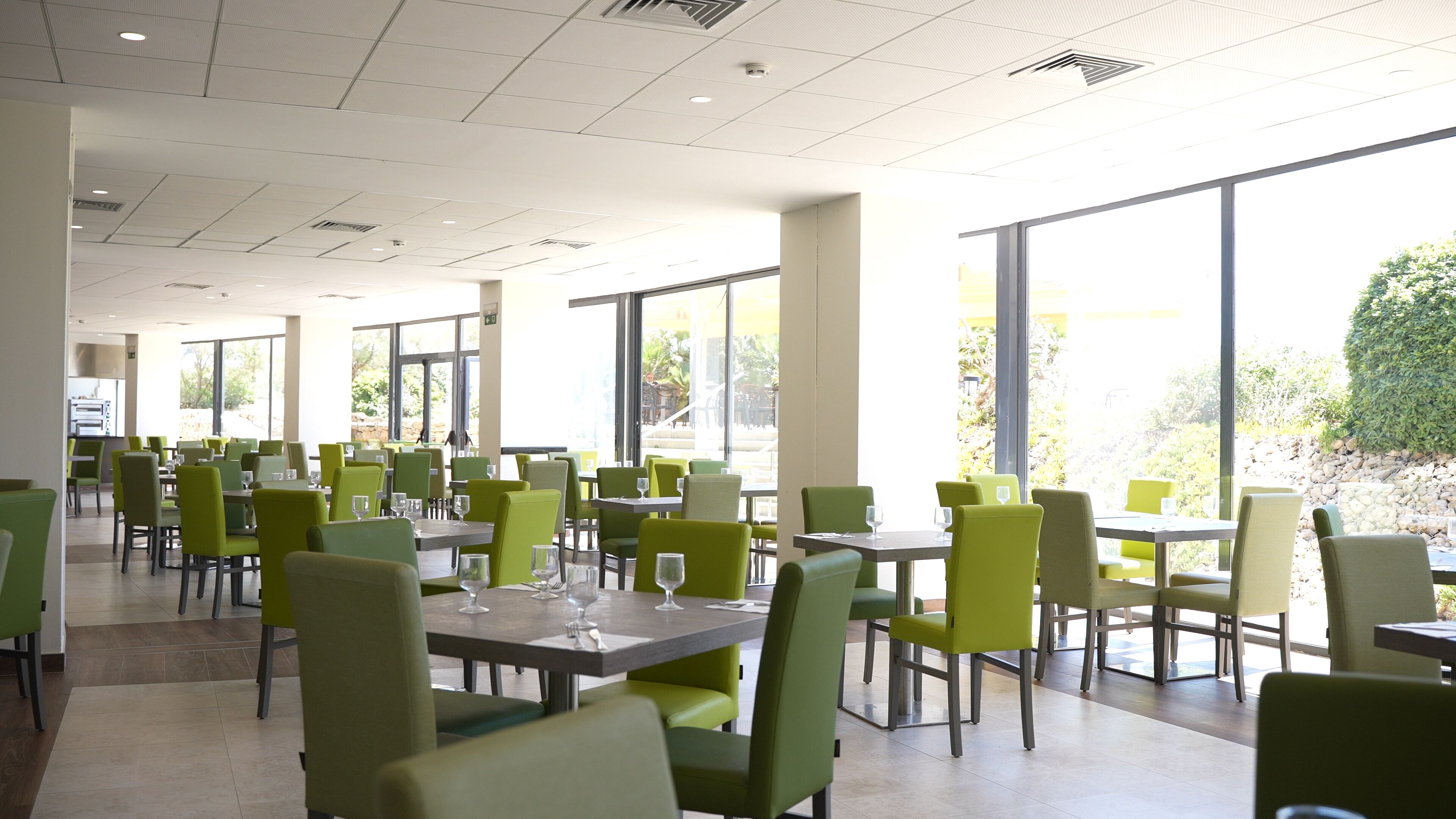 A spacious dining area with green chairs and tables, large windows offering natural light, and a clean, modern ceiling design.