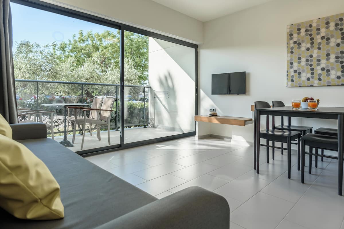 A modern living room with a sofa, a dining area with a table and chairs, a flat-screen TV, and a large window offering a view of the outdoors.
