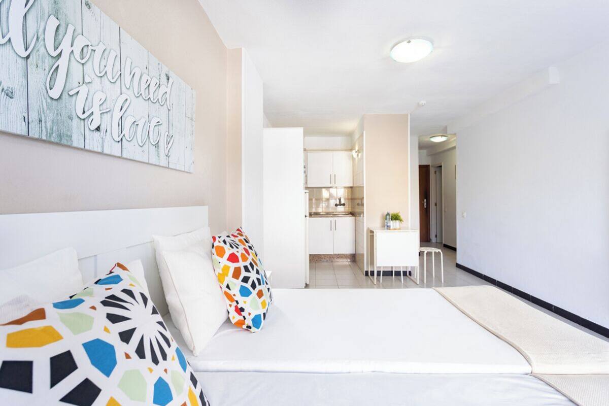 Moderni hotellihuone, jossa on sänky, värikkäitä tyynyjä, taustalla keittiö ja käytävä, joka johtaa toiseen huoneeseen.