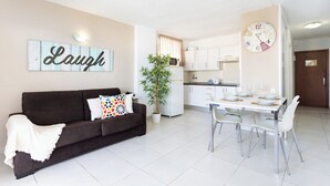 Un salon moderne avec un canapé marron foncé, une table à manger avec des chaises, un coin cuisine et une horloge au mur.