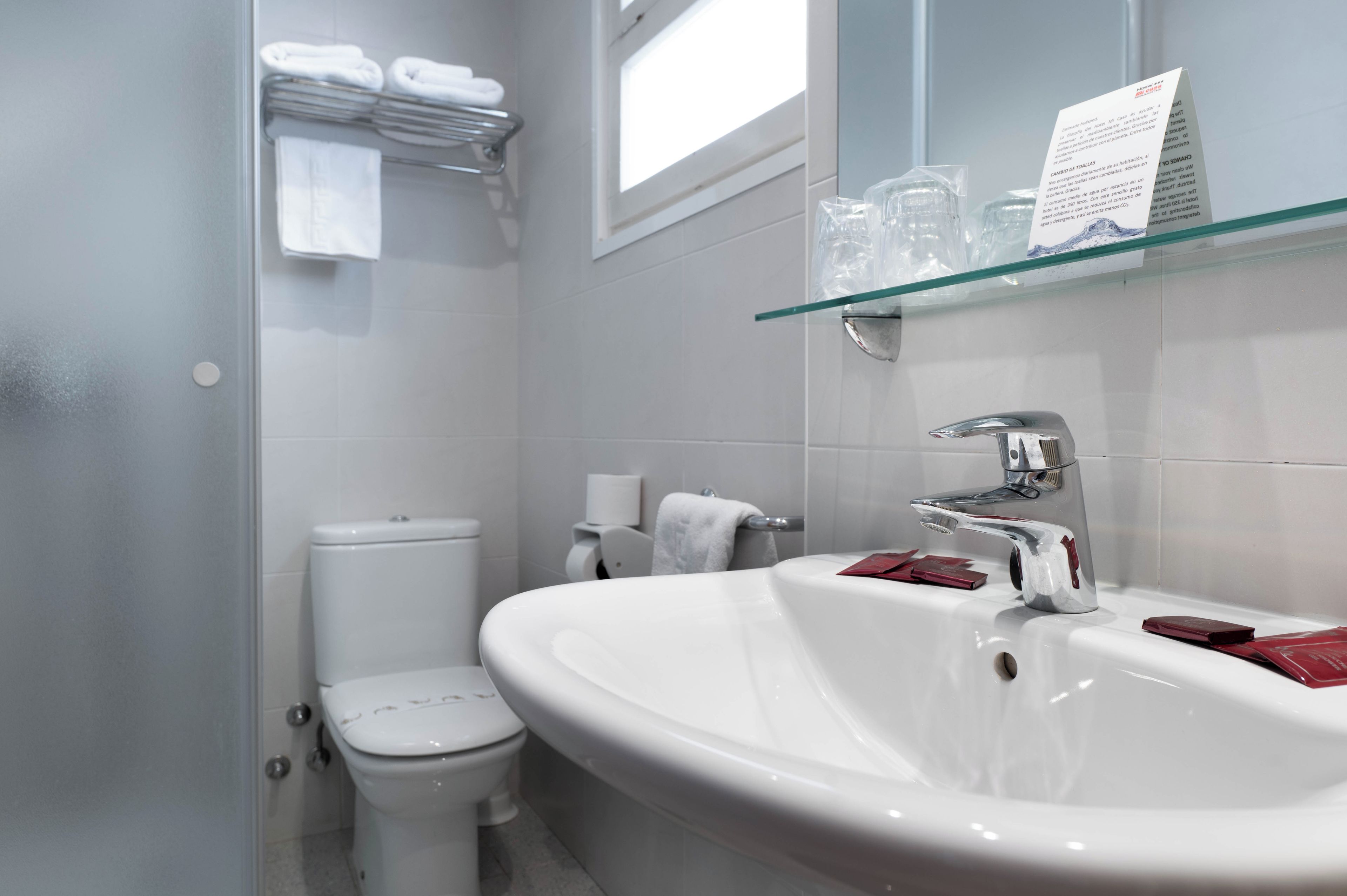 A modern bathroom with a white toilet, a large white sink, and a glass shelf above the sink.