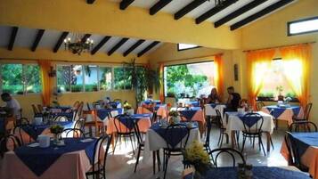 Ein Restaurant mit mehreren Tischen und Stühlen, großen Fenstern mit Vorhängen und einer Decke mit freiliegenden Balken.