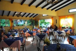 Un ristorante con numerosi tavoli e sedie, ampie finestre con tende e un soffitto con travi a vista.