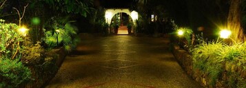 A pathway leading to a structure with a roof, surrounded by greenery and illuminated by lights.
