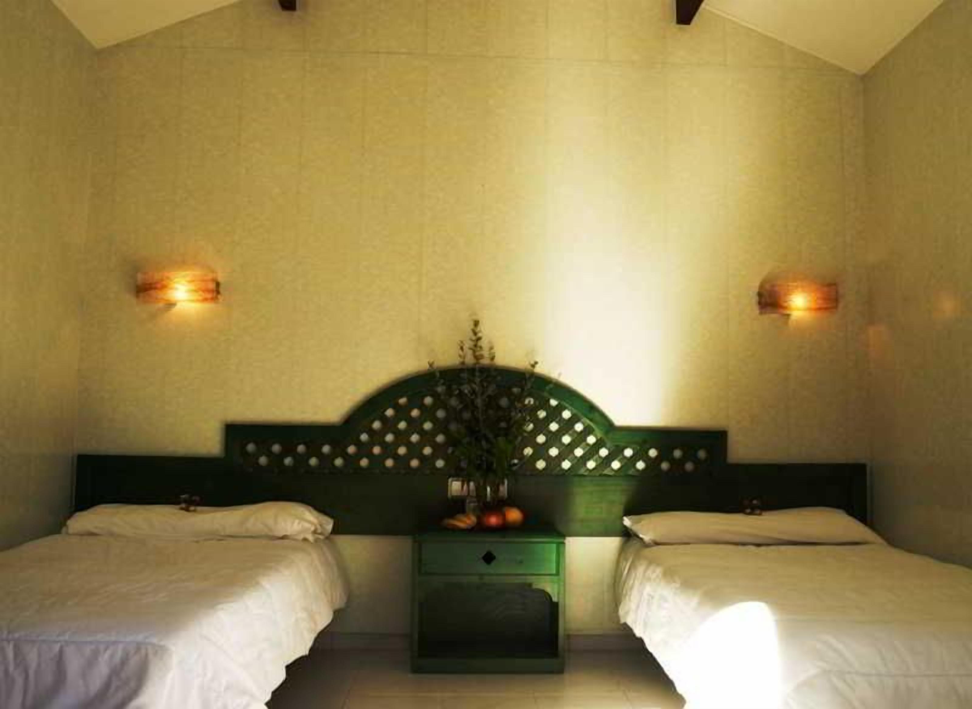 Ein Hotelzimmer mit zwei Einzelbetten, einem grünen Nachttisch mit Blumenvase und Wandleuchten.