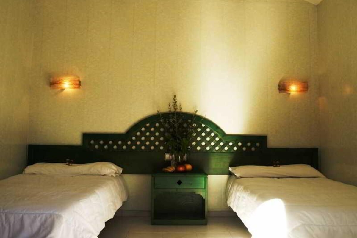 Une chambre d’hôtel avec deux lits simples, une table de chevet verte ornée d’un vase de fleurs et des lampes fixées au mur.