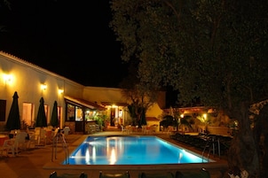 Piscina com espreguiçadeiras e guarda-sóis, cercada por um edifício com área de estar ao ar livre e bar.