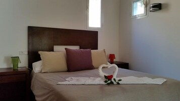 A hotel room with a bed, a nightstand, a green vase, and a heart-shaped towel decoration.
