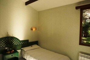 Quarto pequeno com cama de solteiro, cabeceira verde, mesa de cabeceira com uma planta e janela com vista para vegetação.