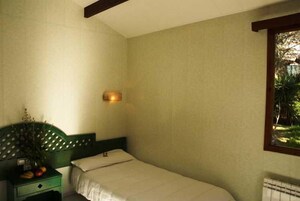 A small, single-bed room with a green headboard, a bedside table with a plant, and a window with a view of greenery.