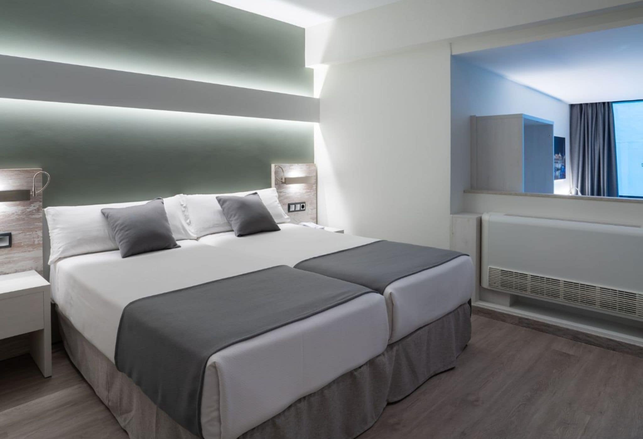 A modern hotel room with a large bed, bedside tables, a built-in TV, and a view of the city.