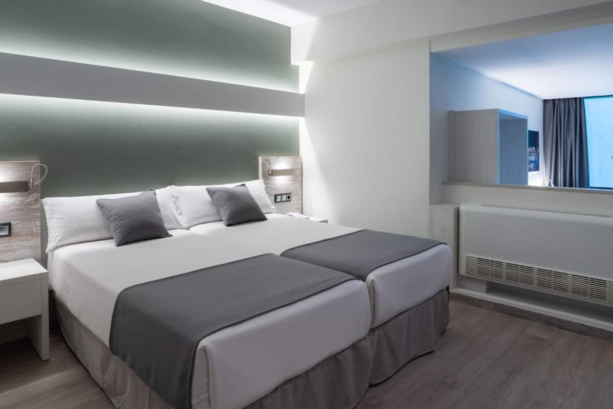 A modern hotel room with a large bed, bedside tables, a built-in TV, and a view of the city.