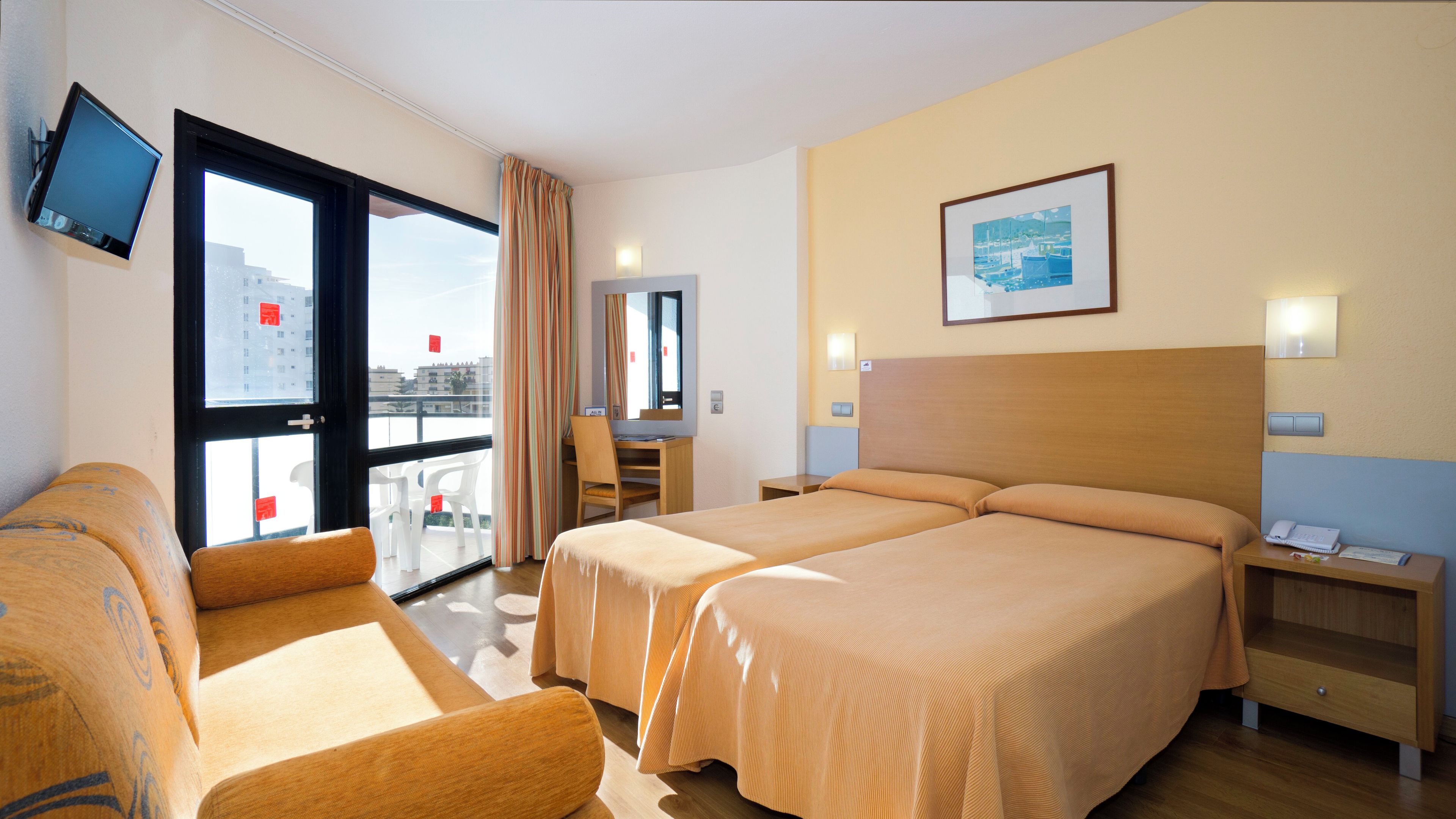 A hotel room with two beds, a desk, a chair, a TV, and a balcony with a view of buildings.