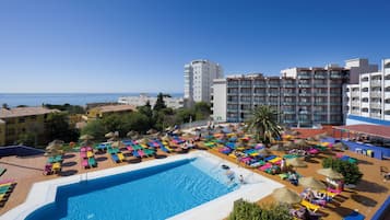 A pool area with multiple lounge chairs and umbrellas, surrounded by buildings and a clear sky.