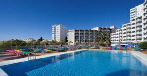 Una piscina con sillas de descanso y sombrillas de colores, rodeada de edificios de varios pisos.