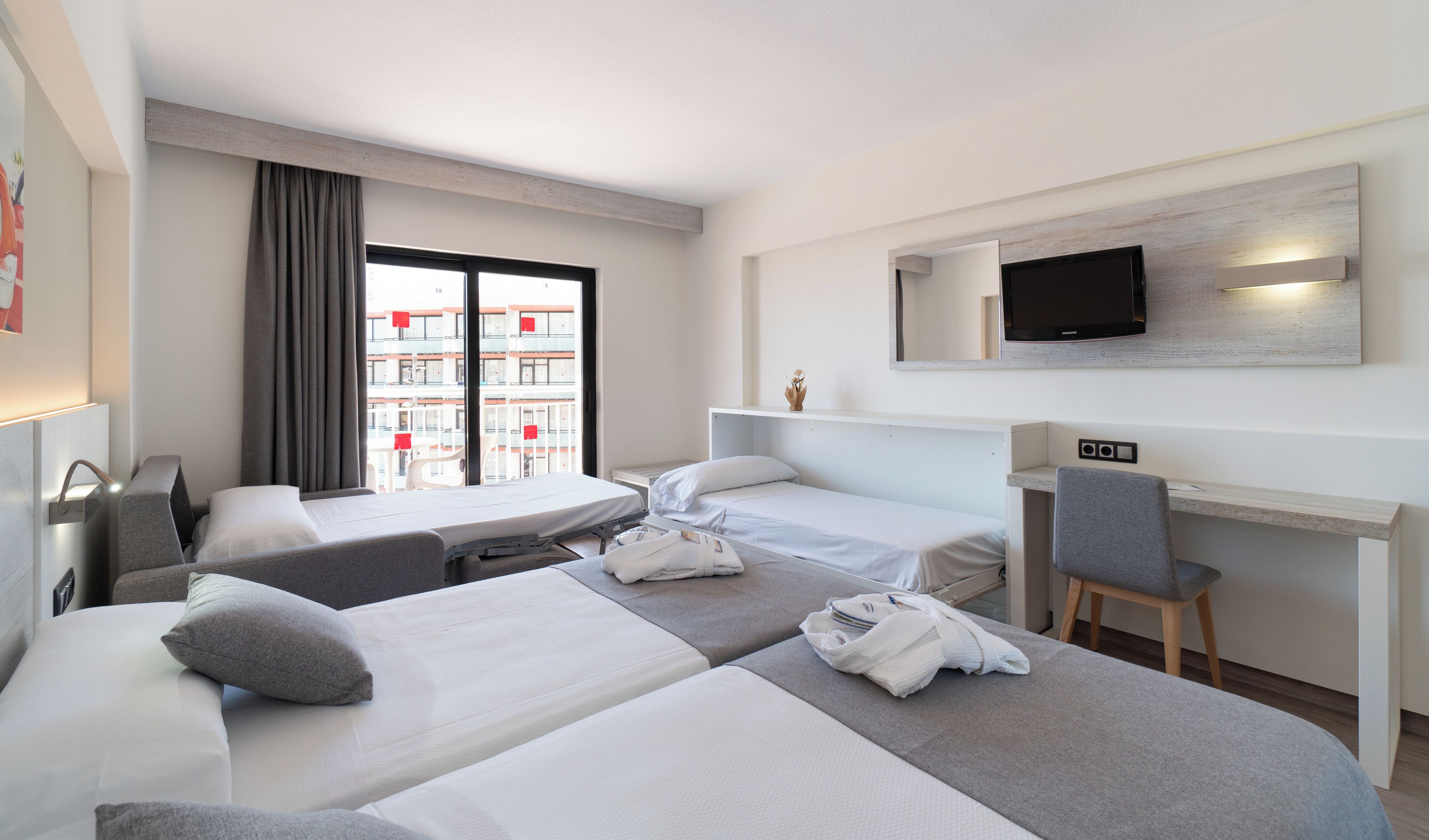 Habitación de hotel con dos camas, un escritorio, una silla, un televisor y ventana con vistas a edificios.