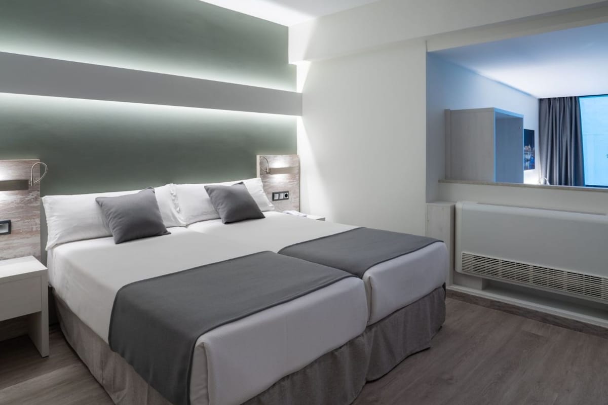 A modern hotel room with a large bed, bedside tables, a built-in TV, and a view of the city.