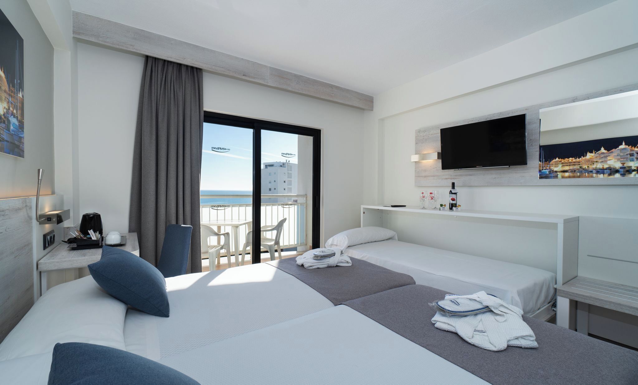 A hotel room with two beds, a desk with a chair, a TV, and a balcony with a view of the city.