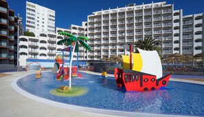 A swimming pool with a ship-shaped play structure, surrounded by a fence and palm trees. - Medplaya Bali (Benalmádena)