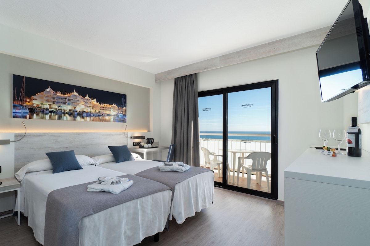A hotel room with two beds, a desk, a chair, a TV, and a balcony with a view of the sea.