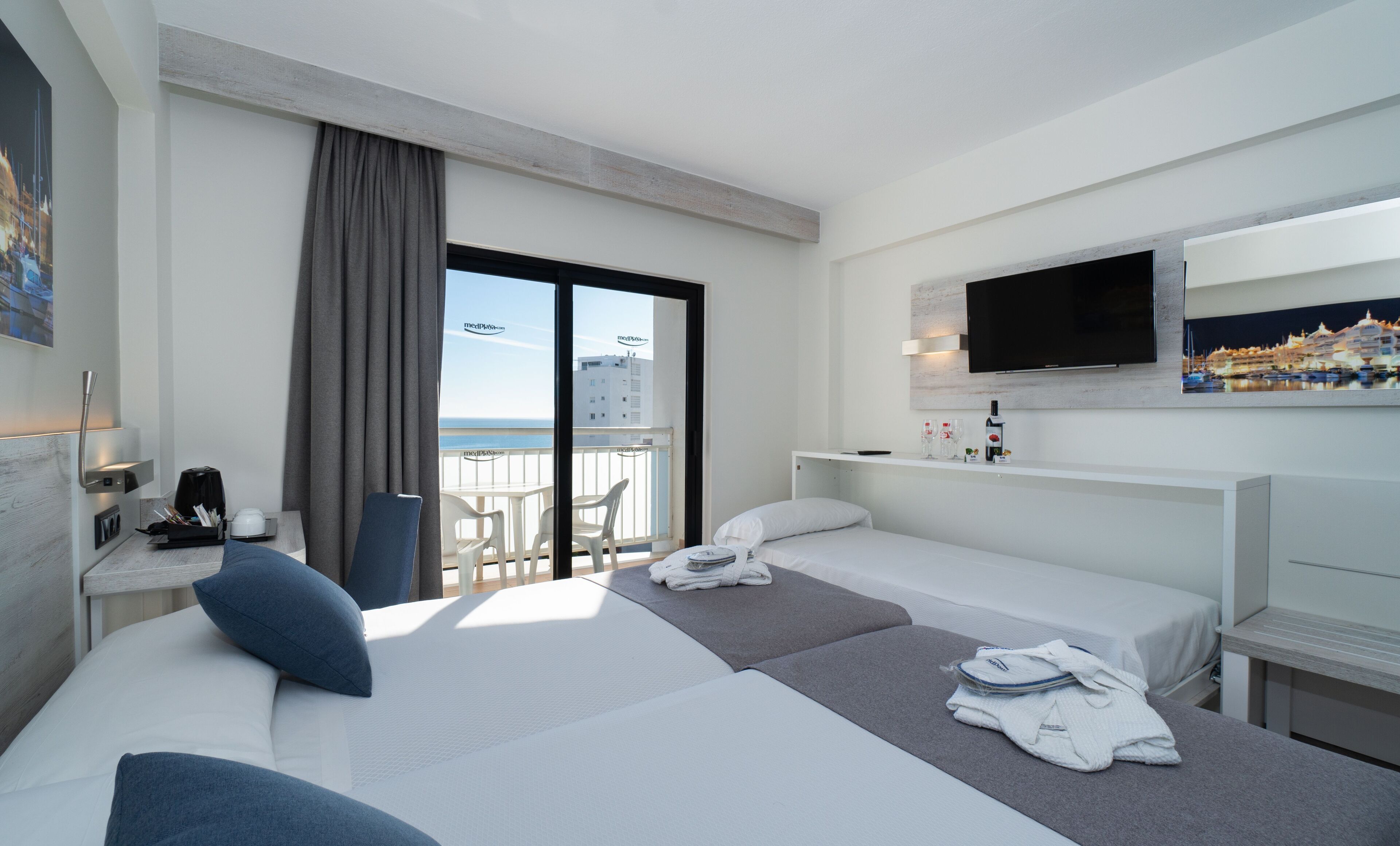 A hotel room with two beds, a desk with a computer, a chair, a TV, and a balcony with a view of the city.