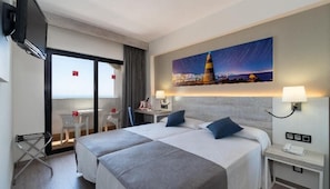 Habitación de hotel con cama, dos mesitas de noche, un escritorio, una silla, televisión y un amplio ventanal con vista a la ciudad iluminada de noche.