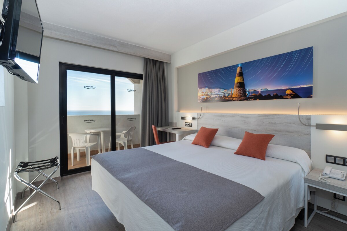 Ett hotellrum med en säng, ett skrivbord, en stol, en telefon och en tavla som föreställer en stadsbild.