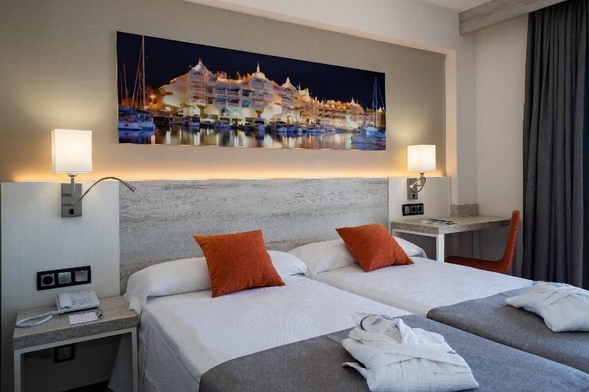 Habitación de hotel con dos camas, una mesita de noche, un escritorio y un cuadro de un paisaje costero.