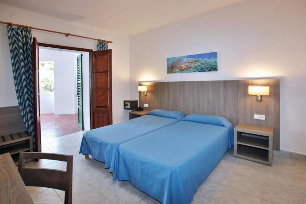 A room with two beds, a wooden headboard, a painting, two bedside lamps, a small table, and a bench.