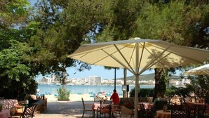 An outdoor dining area with tables and chairs under umbrellas, overlooking a beach and water.