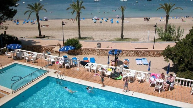 Un área de piscina con sillas de descanso y sombrillas, una playa con palmeras y barcos al fondo.