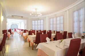 A restaurant with tables set for dining, a chandelier, and large windows with curtains.