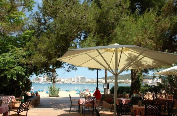 An outdoor seating area with tables and chairs, a large umbrella, and a view of a body of water with buildings in the background. - Hotel Ses Savines (Sant Antoni de Portmany)