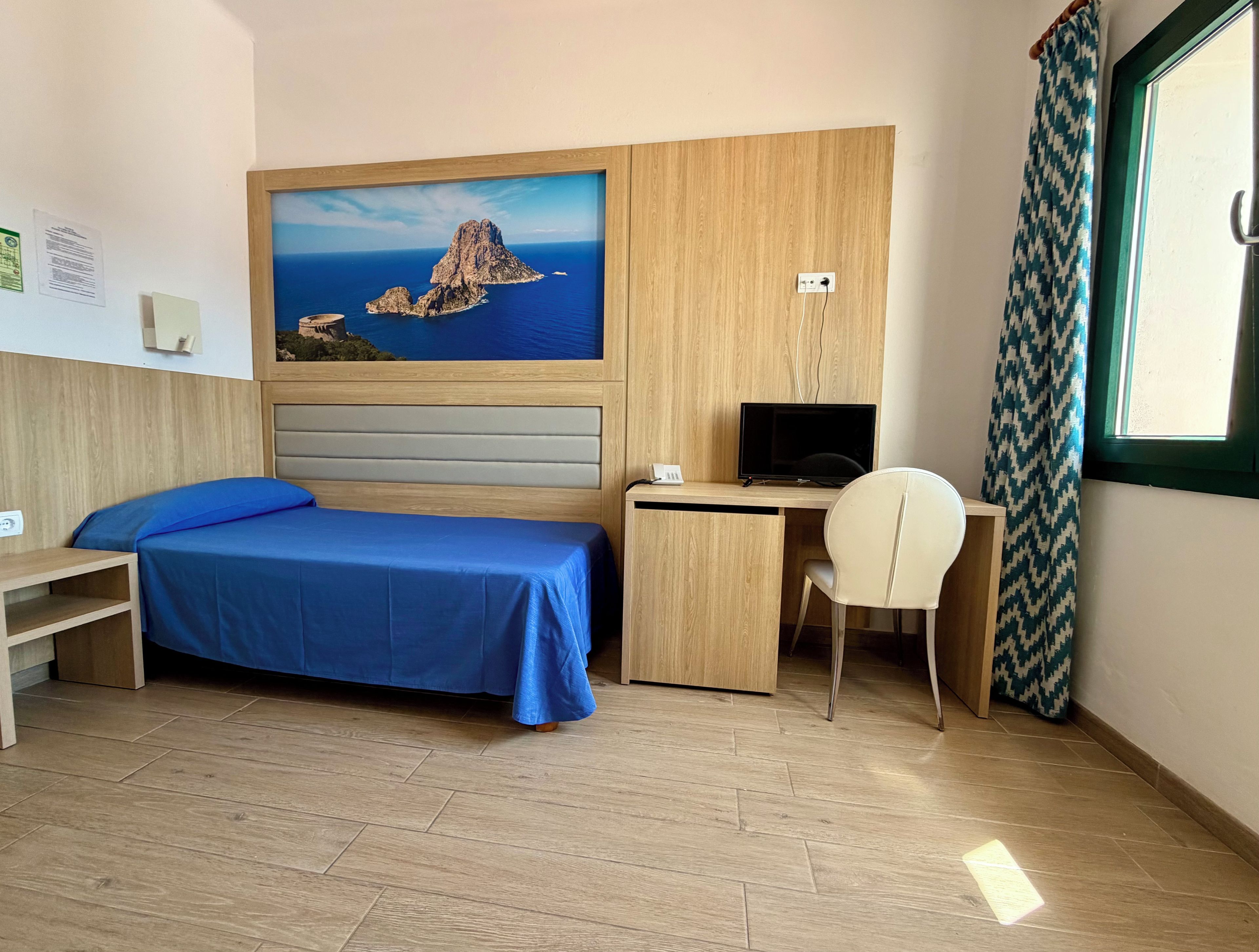 Quarto de hotel com cama, escrivaninha, cadeira e pintura de paisagem costeira.