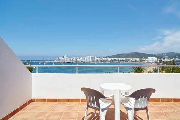 Un balcon avec deux chaises blanches et une petite table, donnant sur une marina où sont amarrés des bateaux, et sur le paysage urbain de la côte.