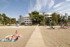 Una playa con un paseo de madera que lleva a un edificio con varios balcones, rodeado de palmeras.