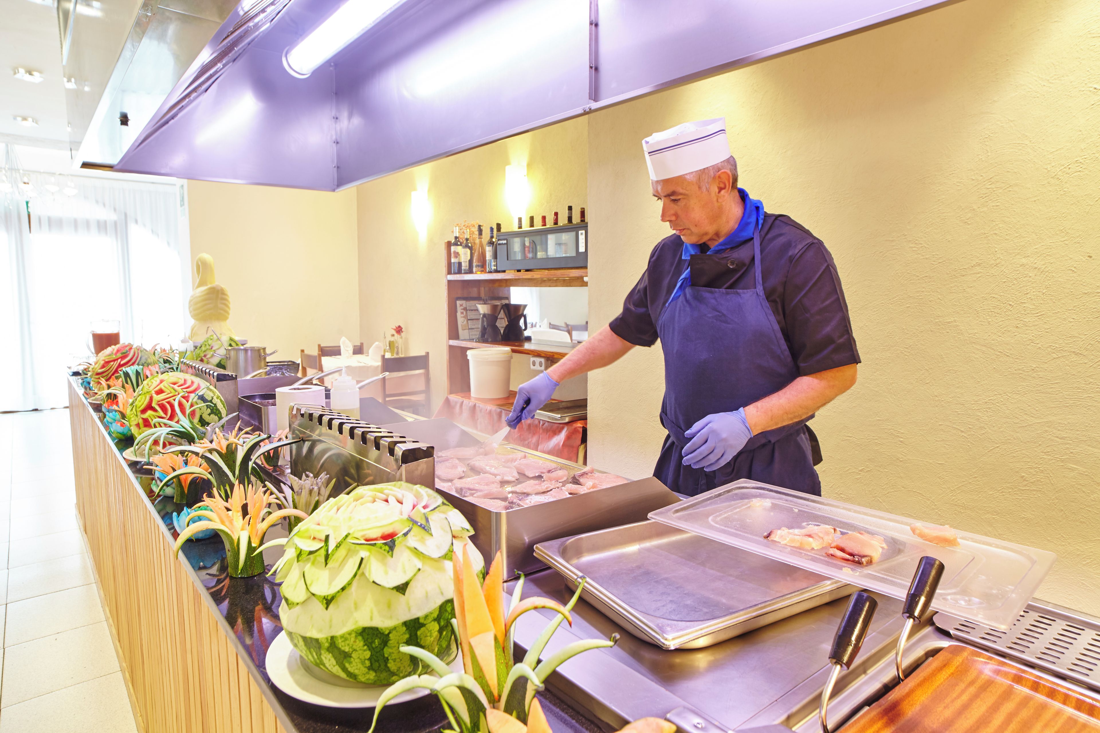 Disposição de buffet com diversos alimentos, incluindo uma melancia esculpida em formato decorativo, e uma pessoa servindo.