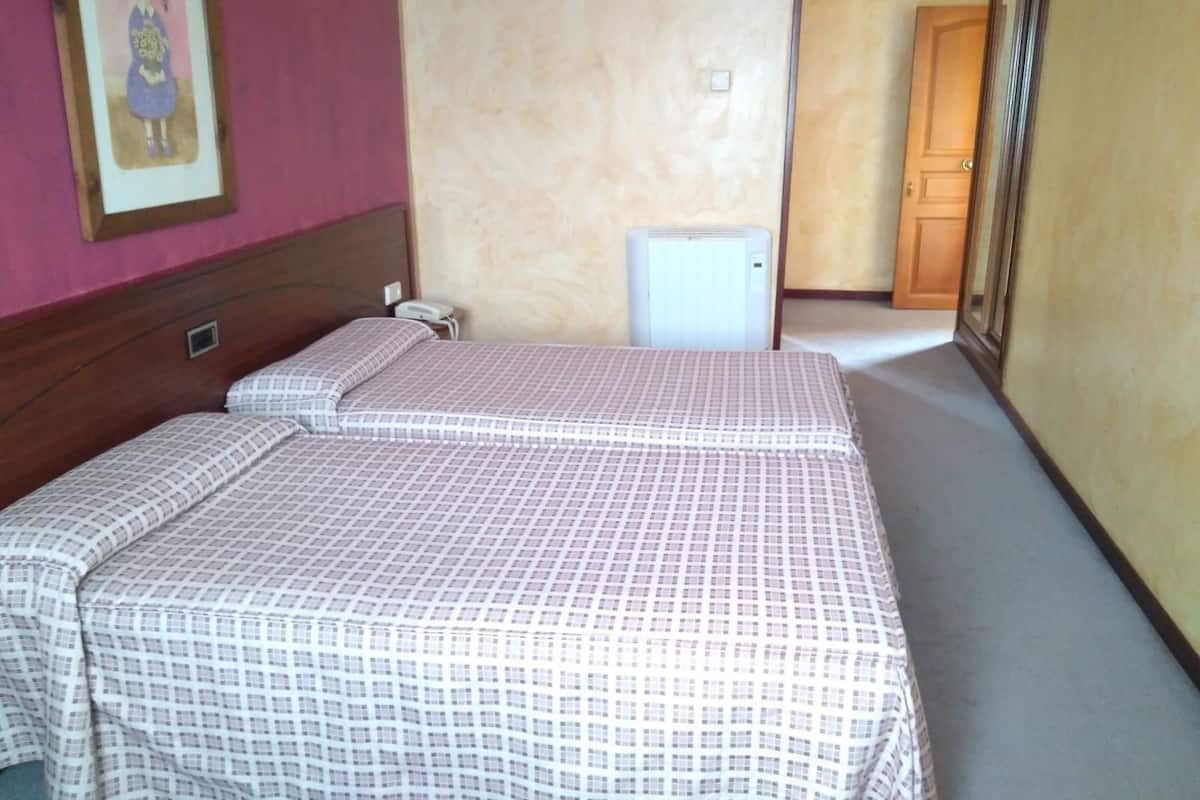 A hotel room with two beds, a small bedside table, a wall-mounted light, and a door leading to another room.