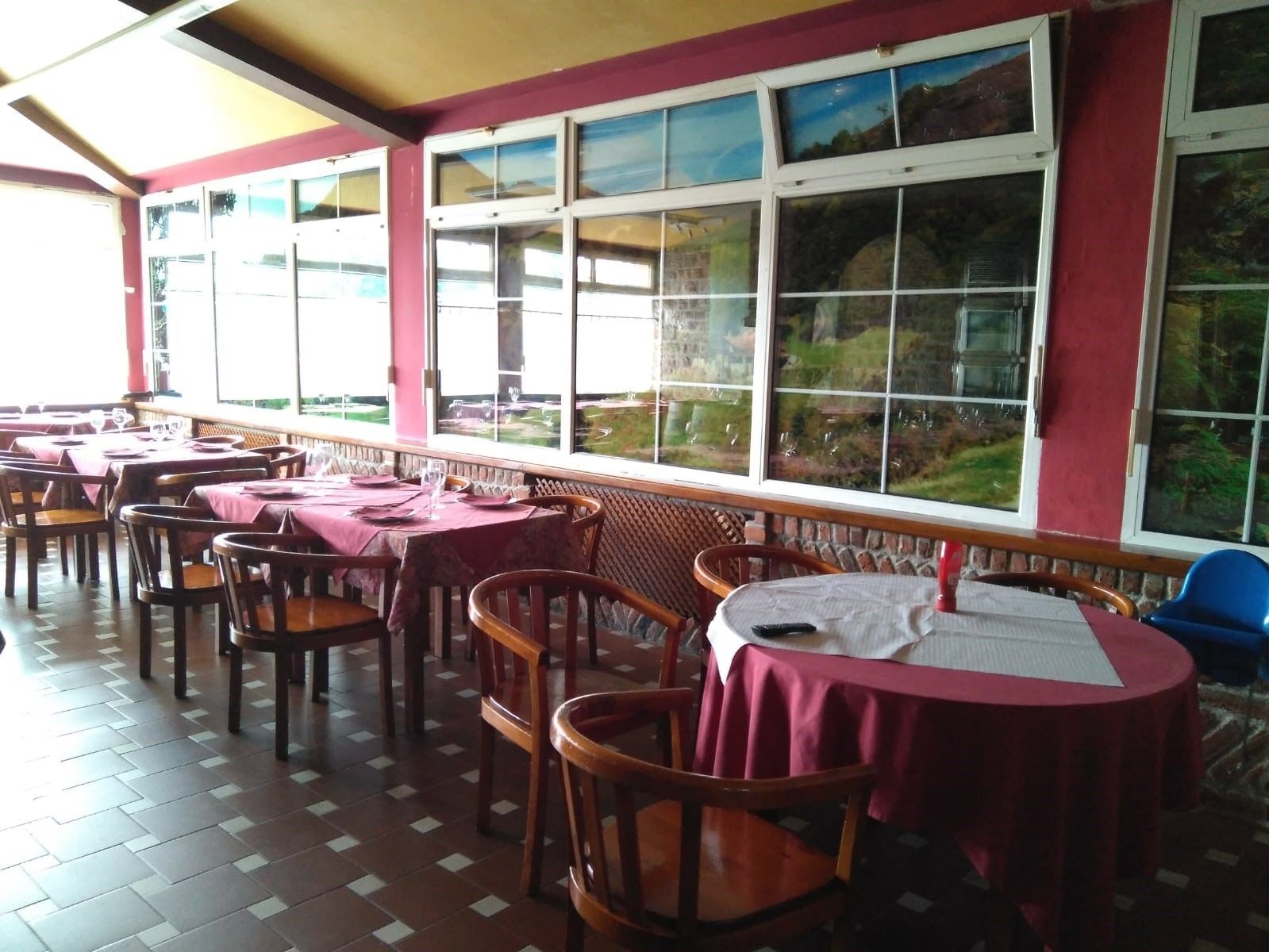 Un restaurante con mesas y sillas, ventanas amplias que dan a zonas verdes y un suelo de baldosas.
