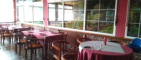 Un restaurante con mesas y sillas, ventanas amplias que dan a zonas verdes y un suelo de baldosas.