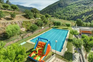 Una piscina con tobogán, rodeada de césped y árboles, con montañas al fondo.