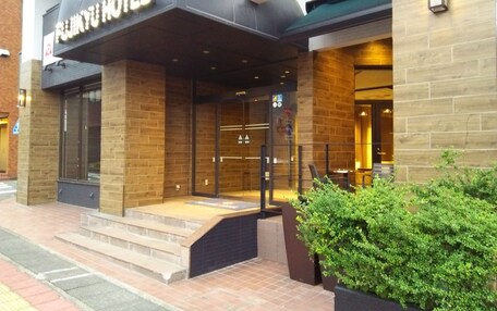 A hotel entrance with a sign reading "FUNNY HOTEL". There are steps leading up to the entrance, a glass door, and a small garden area with bushes to the right.