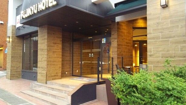 A hotel entrance with a sign reading "FUNNY HOTEL". There are steps leading up to the entrance, a glass door, and a small garden area with bushes to the right. - Fujinomiya Fujikyu Hotel (Fujinomiya)