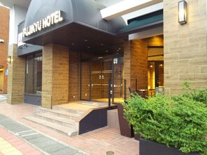 A hotel entrance with a sign reading "FUNNY HOTEL". There are steps leading up to the entrance, a glass door, and a small garden area with bushes to the right. - Fujinomiya Fujikyu Hotel (Fujinomiya)